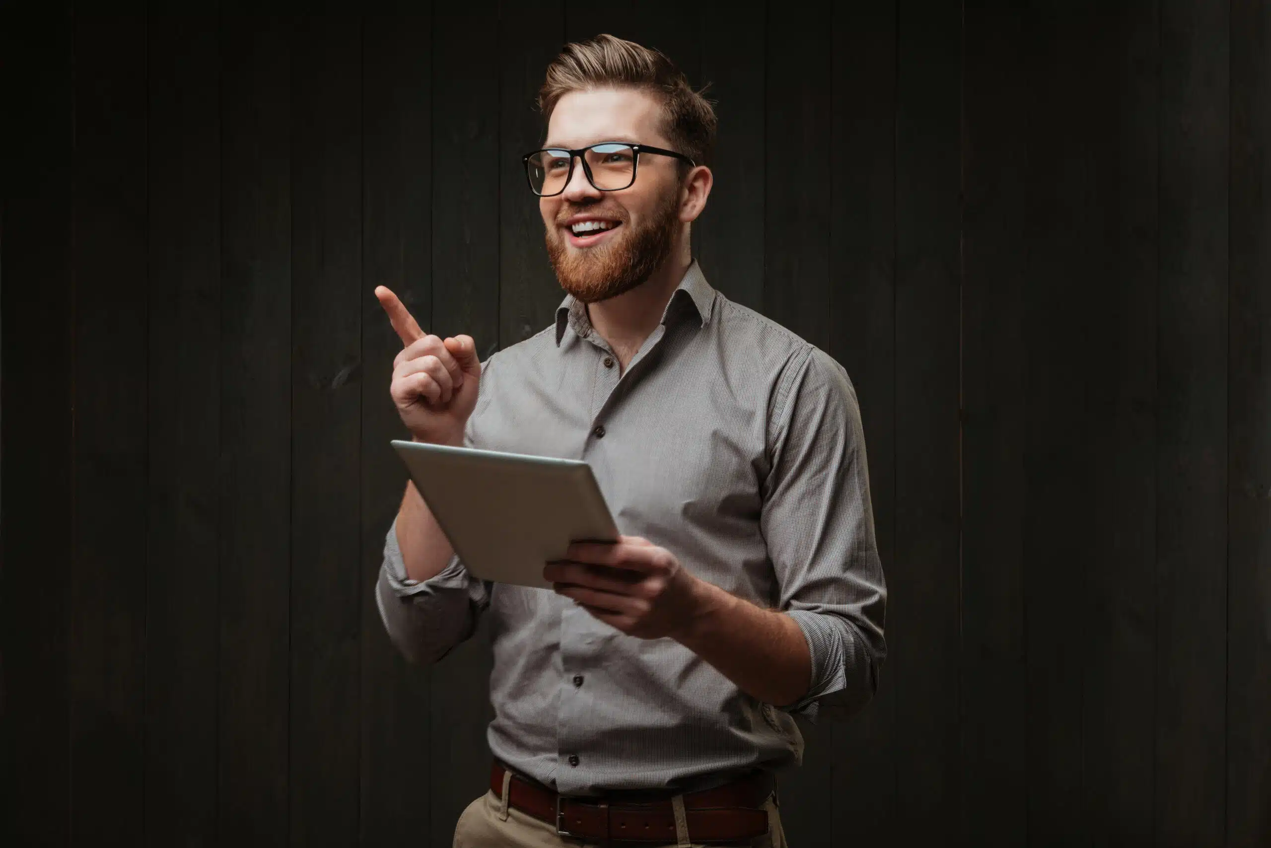 retrato de um homem sorridente e feliz usando oculos segurando um computador tablet e apontando o dedo isolado em uma superficie de madeira preta scaled